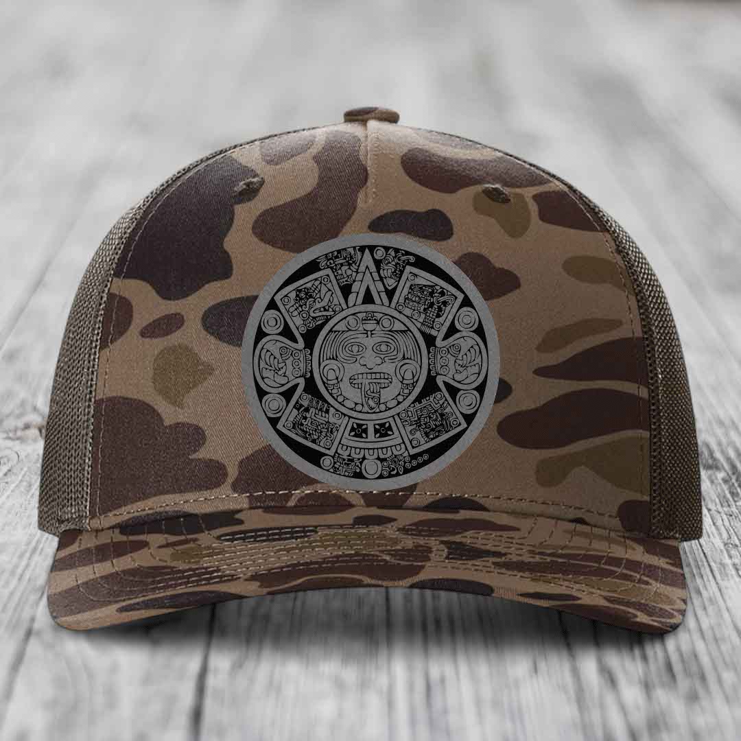 Camouflage trucker hat with Aztec calendar patch on front, placed on wooden surface.