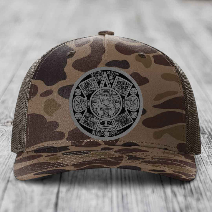 Camouflage trucker hat with Aztec calendar patch on front, placed on wooden surface.