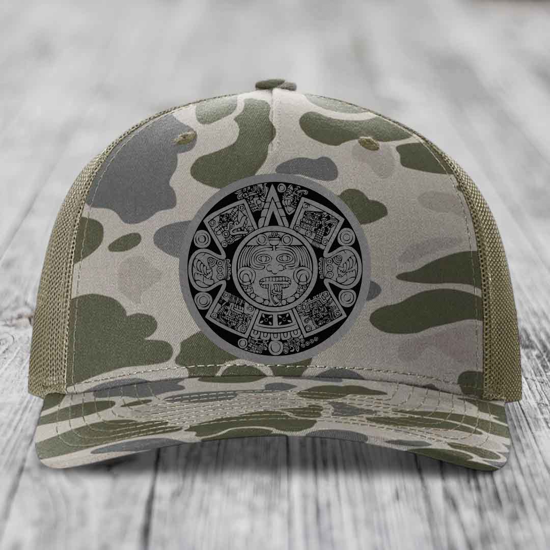 Camouflage trucker hat with Aztec calendar patch on front, placed on wooden surface.