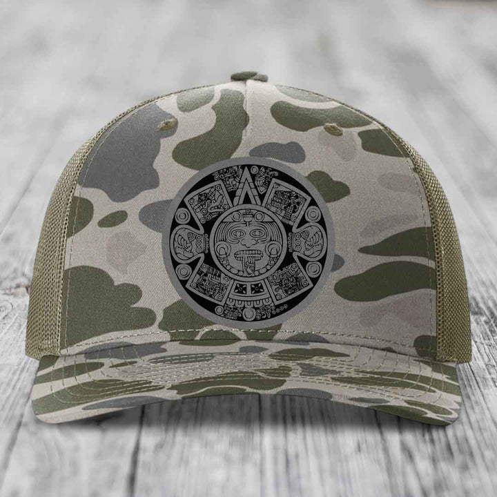 Camouflage trucker hat with Aztec calendar patch on front, placed on wooden surface.