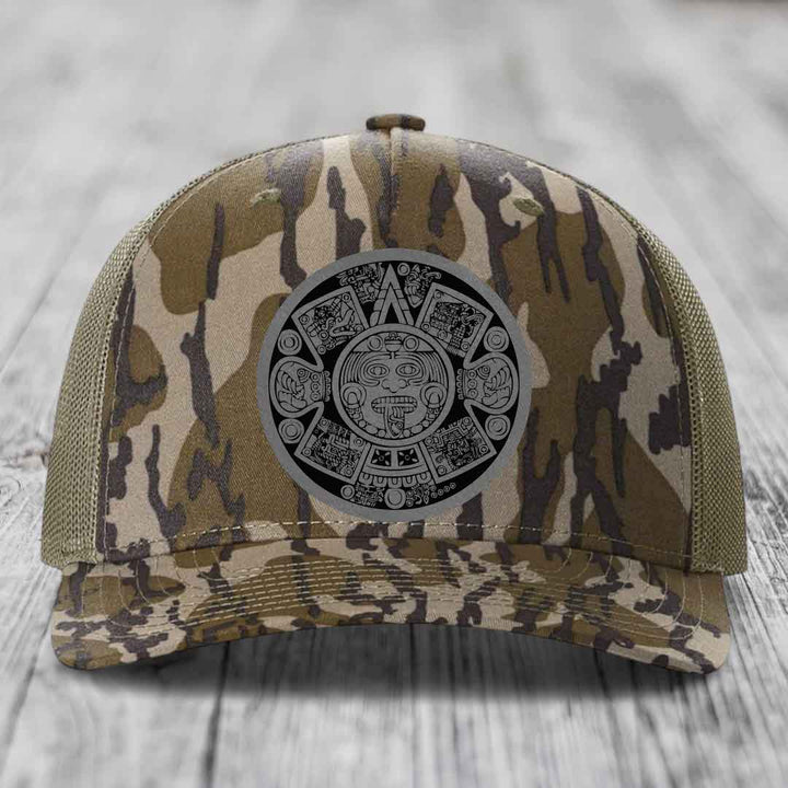 Camouflage trucker hat with Aztec calendar patch on front, displayed on wooden surface.