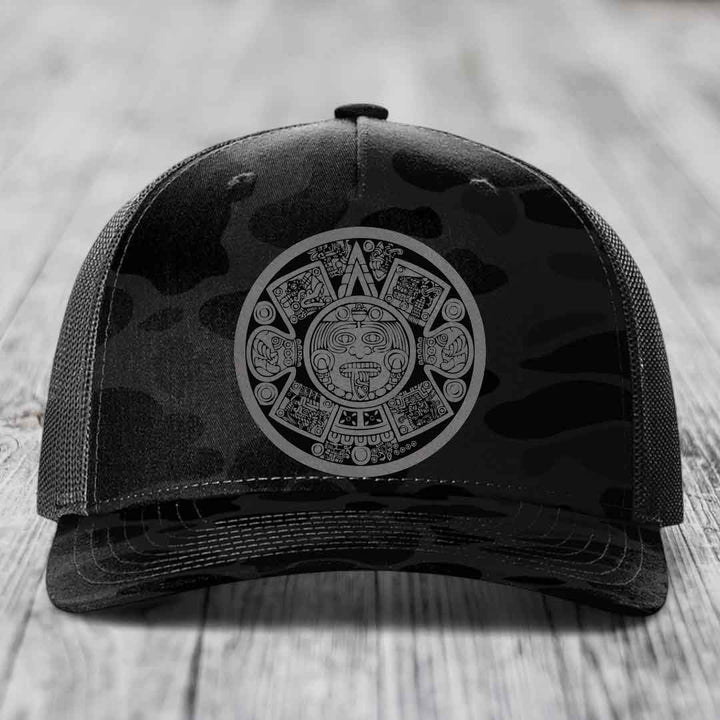 Black camouflage trucker hat with Aztec calendar patch on front.