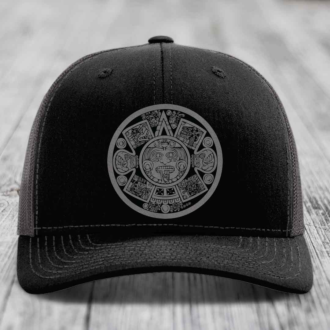 Black trucker hat with grey Aztec calendar patch on front, sitting on wooden surface.