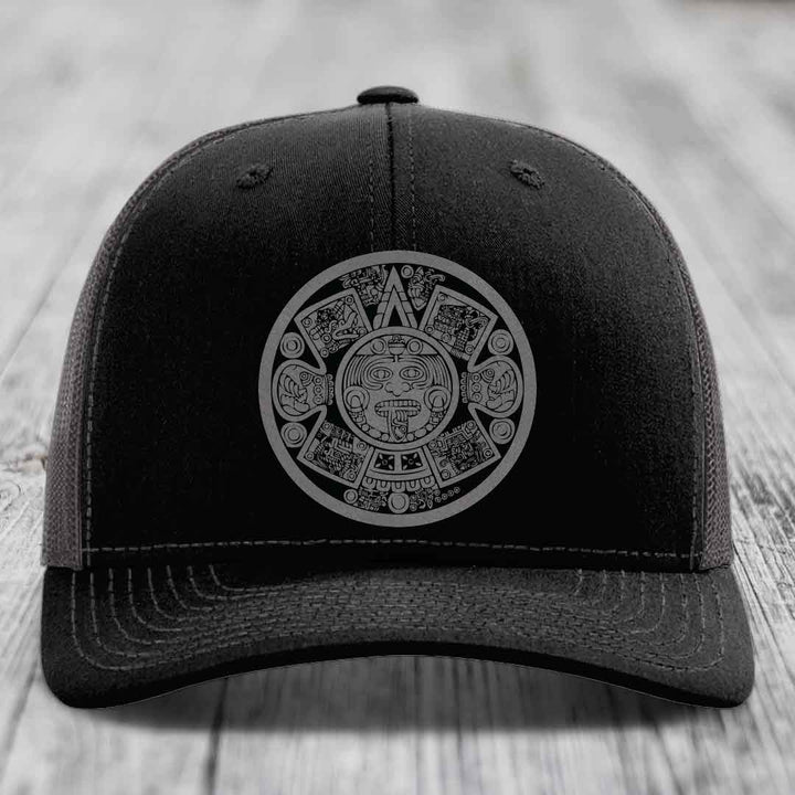 Black trucker hat with grey Aztec calendar patch on front, sitting on wooden surface.