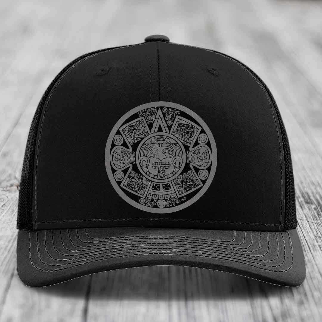 Black trucker hat with grey Aztec calendar patch on front, placed on wooden surface.