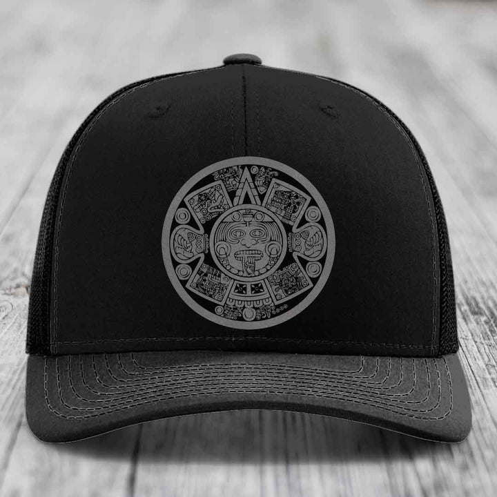 Black trucker hat with grey Aztec calendar patch on front, placed on wooden surface.