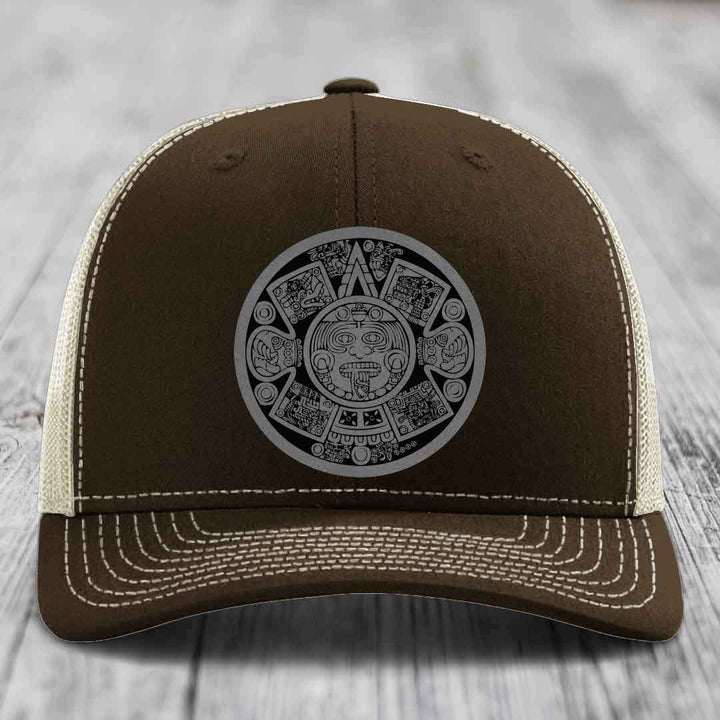 Brown trucker hat with white stitching and a grey Aztec calendar patch on front.
