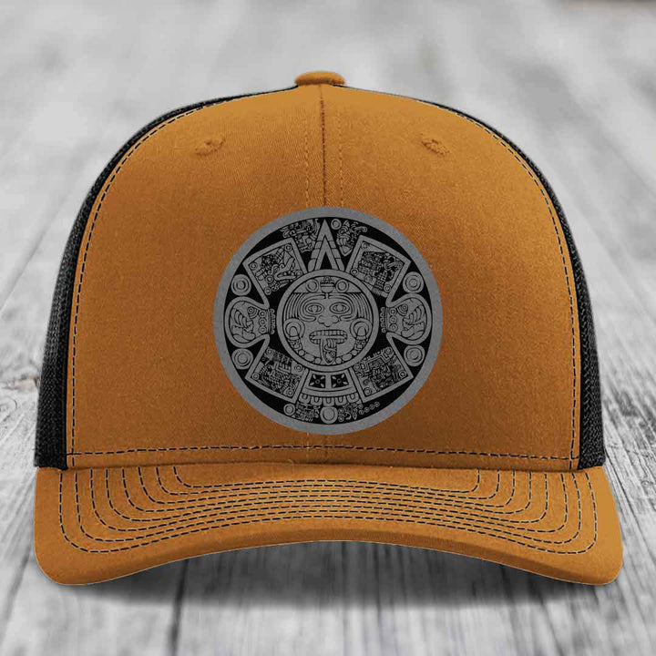 Tan trucker hat with black mesh back and gray Aztec calendar patch on front.