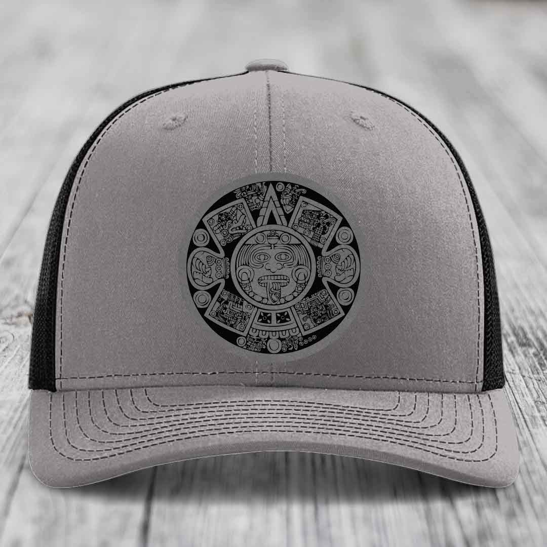 Grey trucker hat with black Aztec calendar patch on front, black mesh back.