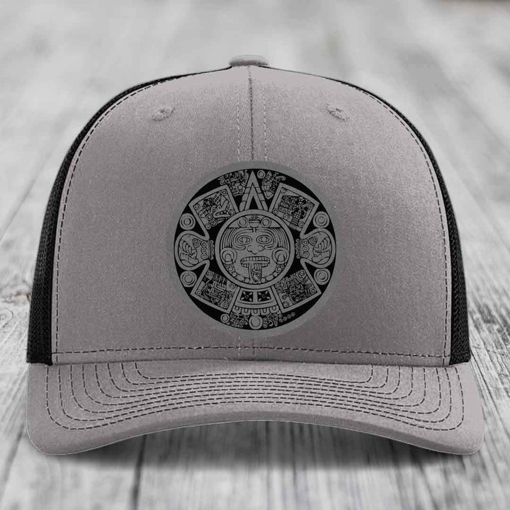 Grey trucker hat with black Aztec calendar patch on front, black mesh back.