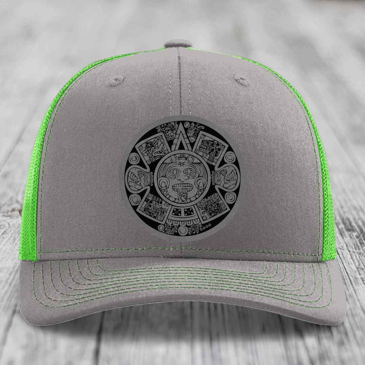 Grey trucker hat with neon green mesh and stitching, featuring a black Aztec calendar patch on the front.