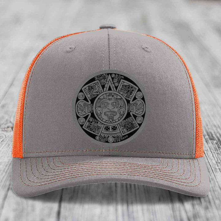 Grey trucker hat with orange mesh back and black Aztec calendar patch on front.