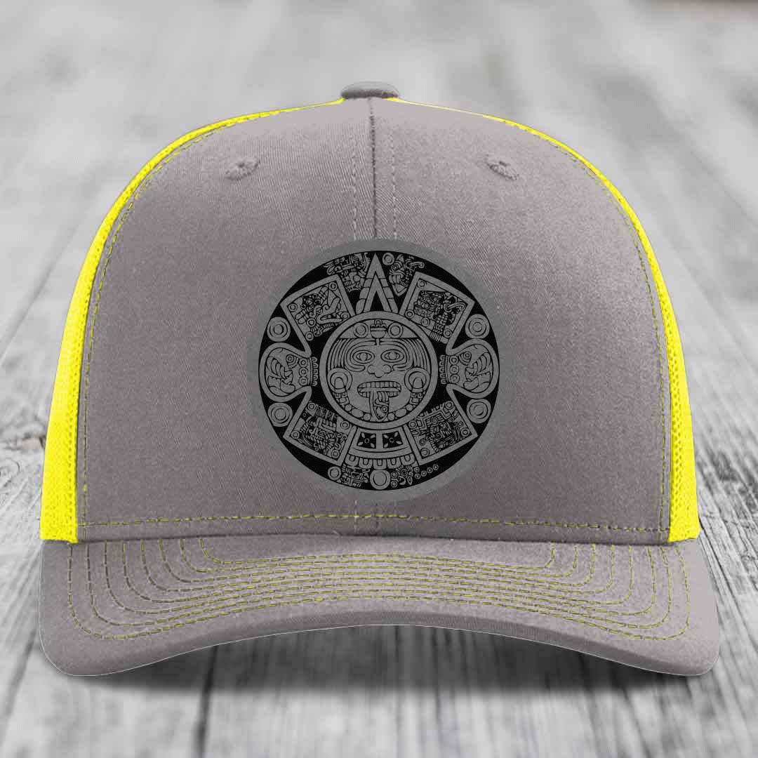 Grey trucker hat with yellow mesh and stitching, featuring a black Aztec calendar patch on front.