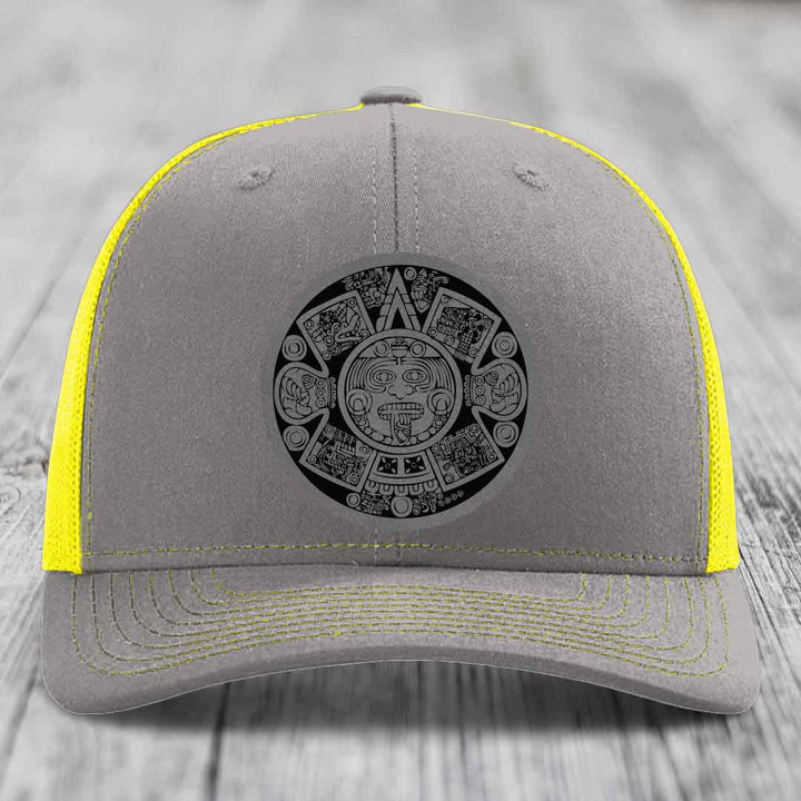 Grey trucker hat with yellow mesh and stitching, featuring a black Aztec calendar patch on front.