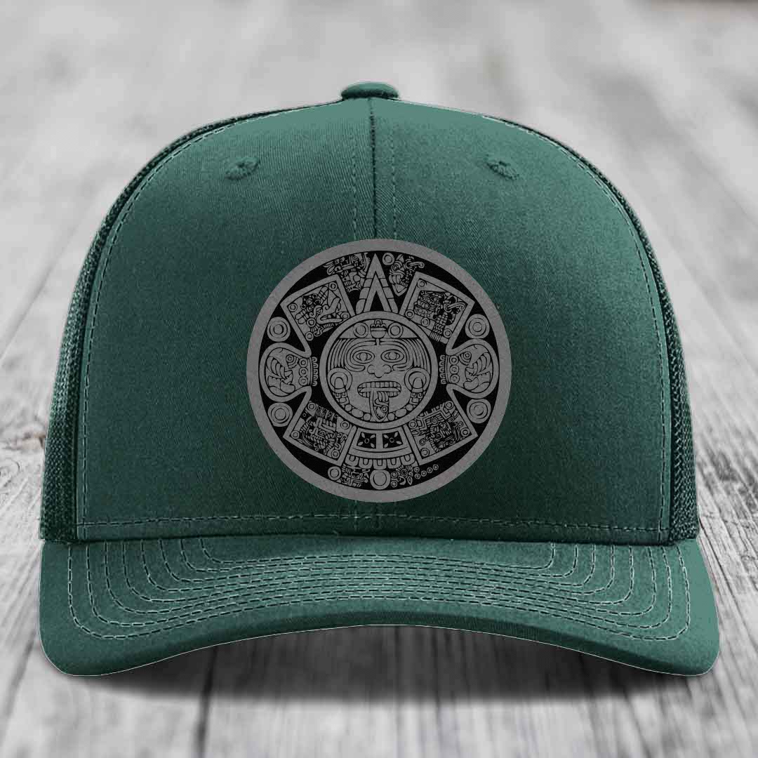 Green trucker hat with Aztec calendar patch on front, displayed on wooden surface.