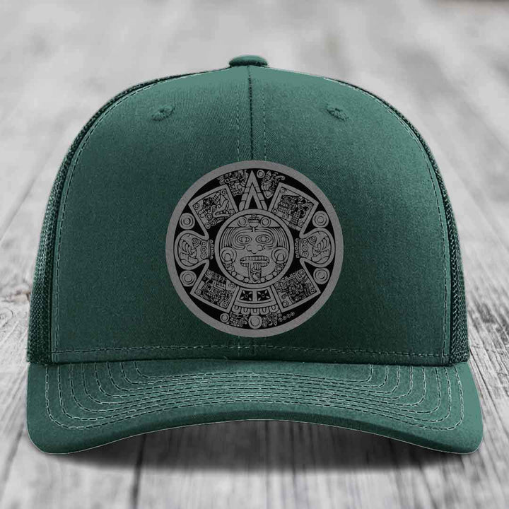 Green trucker hat with Aztec calendar patch on front, displayed on wooden surface.