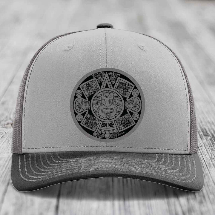 Grey trucker hat with black Aztec calendar patch on front, placed on wooden surface.
