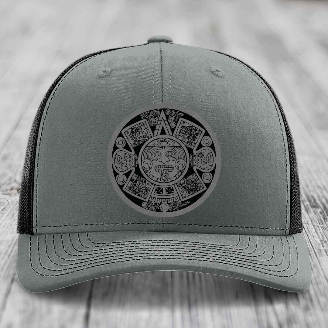 Grey trucker hat with black Aztec calendar patch on front, placed on wooden surface.