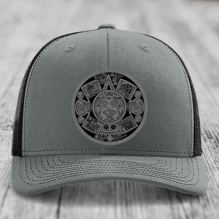 Grey trucker hat with black Aztec calendar patch on front, placed on wooden surface.