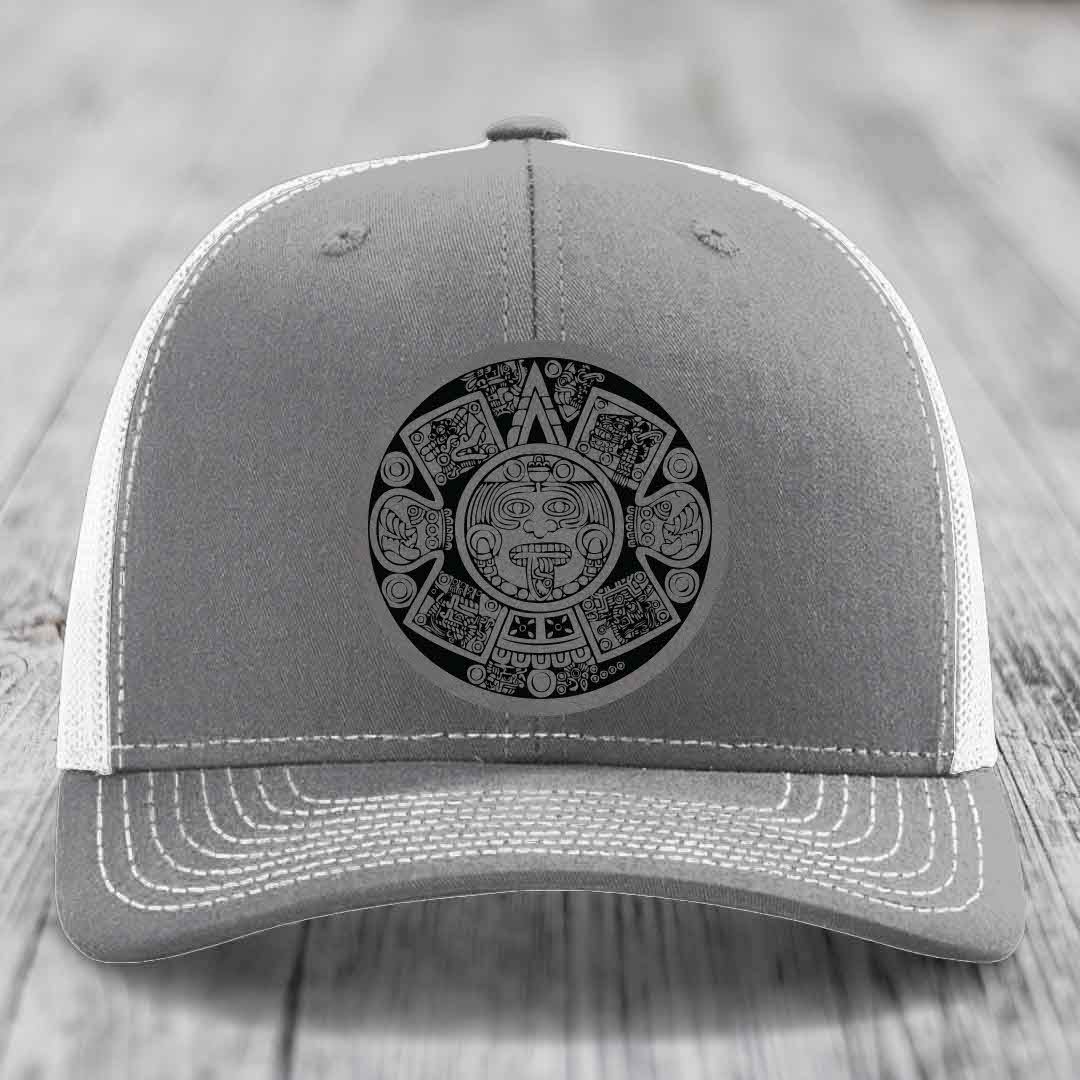 Grey trucker hat with white mesh back and embroidered Aztec calendar design on front.