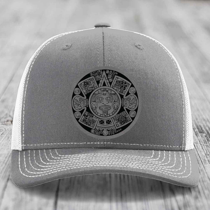 Grey trucker hat with white mesh back and embroidered Aztec calendar design on front.