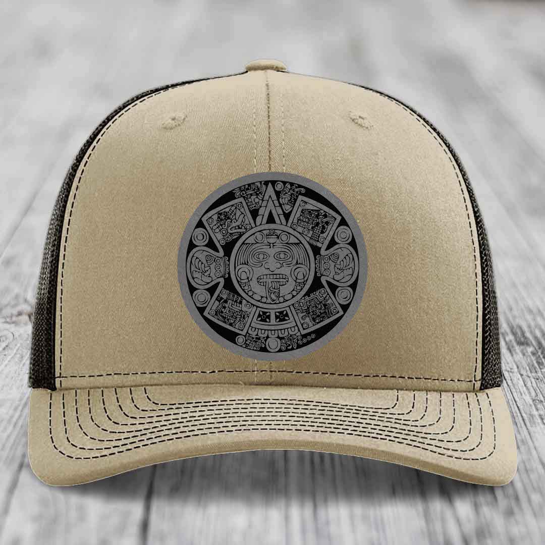 Beige trucker hat with black Aztec calendar patch on front, black mesh back, white stitching.