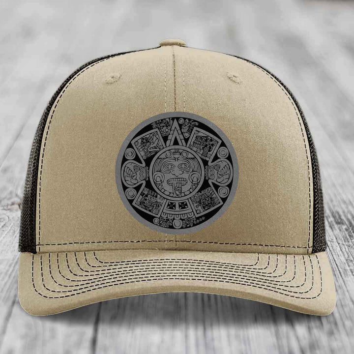 Beige trucker hat with black Aztec calendar patch on front, black mesh back, white stitching.