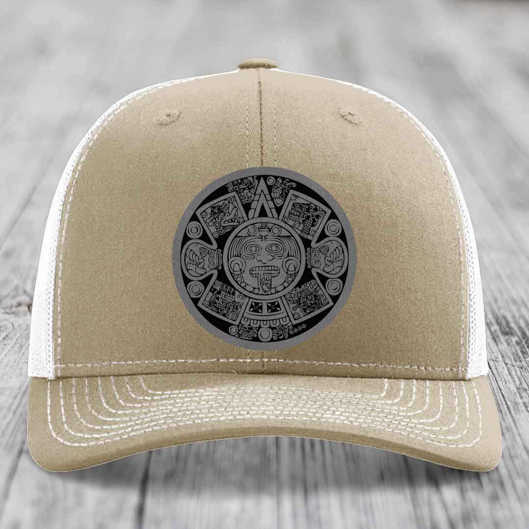 Tan trucker hat with white mesh back and embroidered Aztec calendar patch on front.