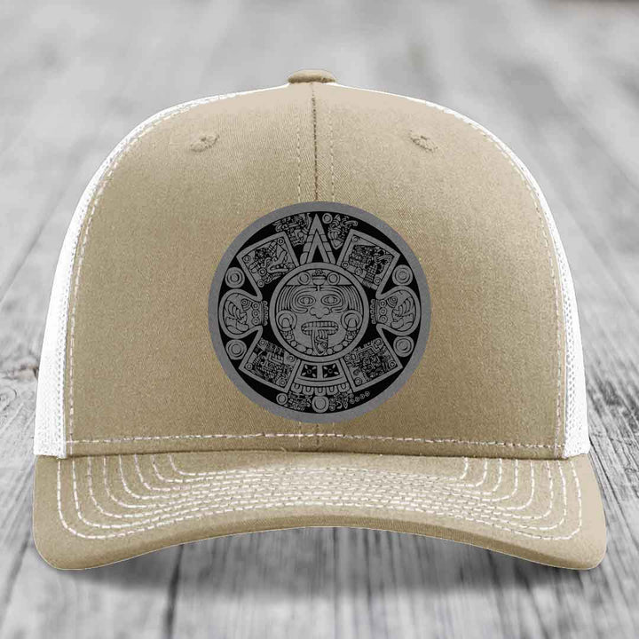 Tan trucker hat with white mesh back and embroidered Aztec calendar patch on front.