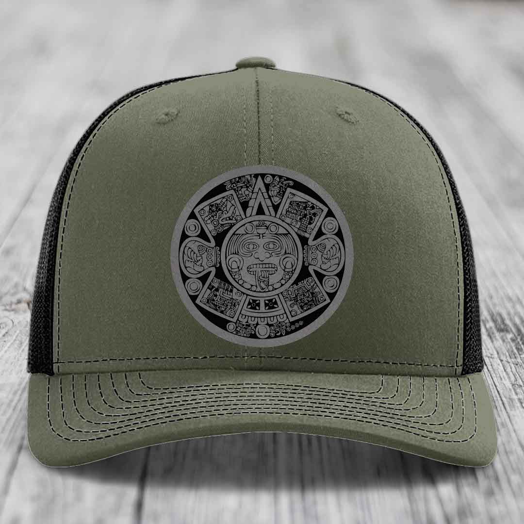 Olive green trucker hat with black mesh back and Aztec calendar patch on front.