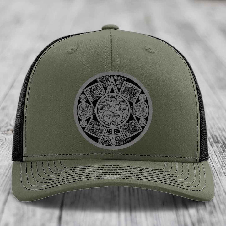 Olive green trucker hat with black mesh back and Aztec calendar patch on front.