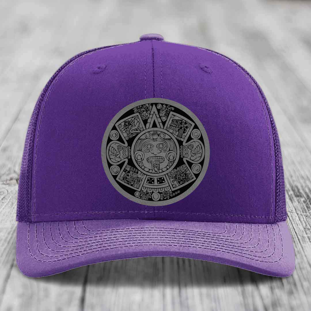 Purple trucker hat with Aztec calendar patch on front, displayed on wooden surface.