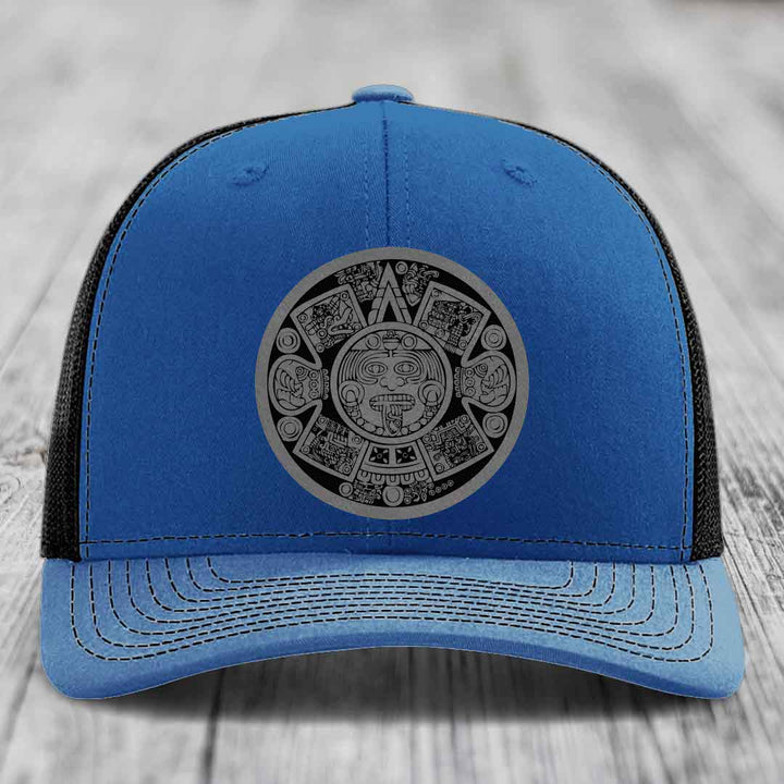 Blue trucker hat with black mesh back and gray Aztec calendar patch on front.