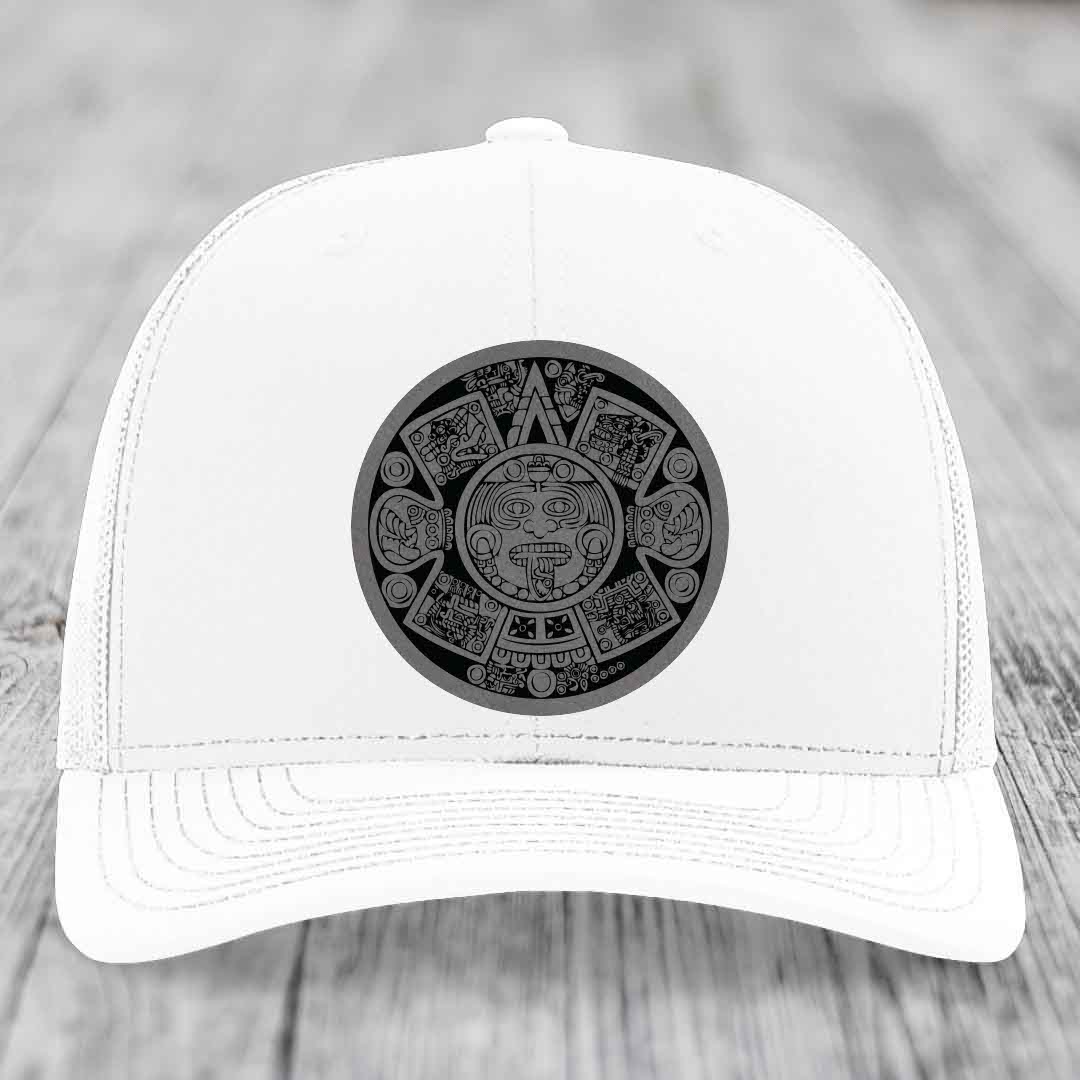 White trucker hat with black Aztec calendar patch on front, placed on wooden surface.
