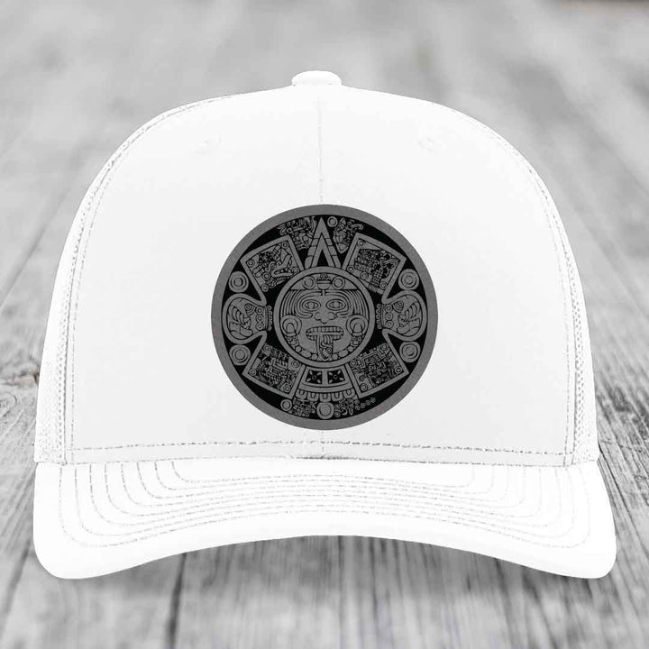 White trucker hat with black Aztec calendar patch on front, placed on wooden surface.