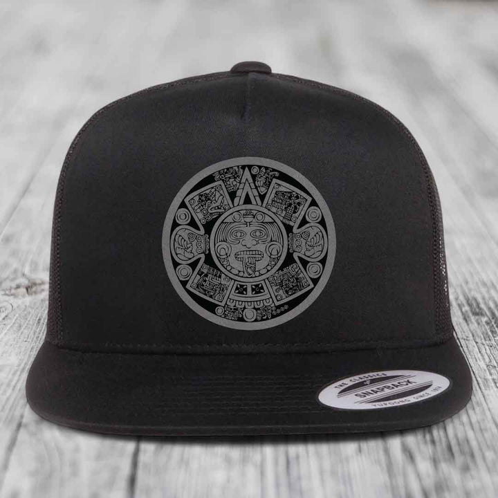 Black snapback hat with Aztec calendar design patch on front, displayed on wooden surface.