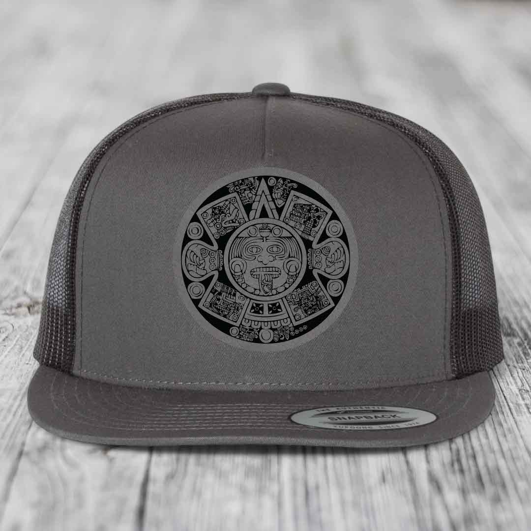 Grey trucker hat with black Aztec calendar patch on front, flat brim, mesh back.