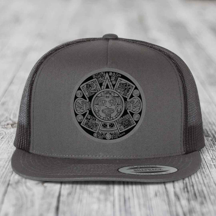 Grey trucker hat with black Aztec calendar patch on front, flat brim, mesh back.