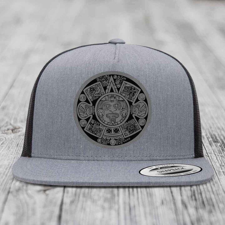 Grey trucker hat with black Aztec calendar patch on front, black mesh back, flat brim.