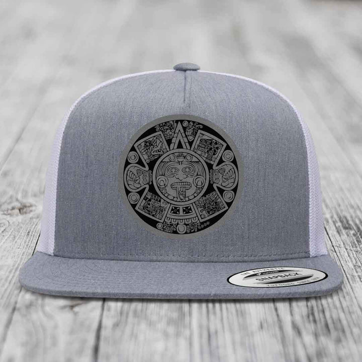 Grey trucker hat with black Aztec calendar patch on front, white mesh back, flat brim.