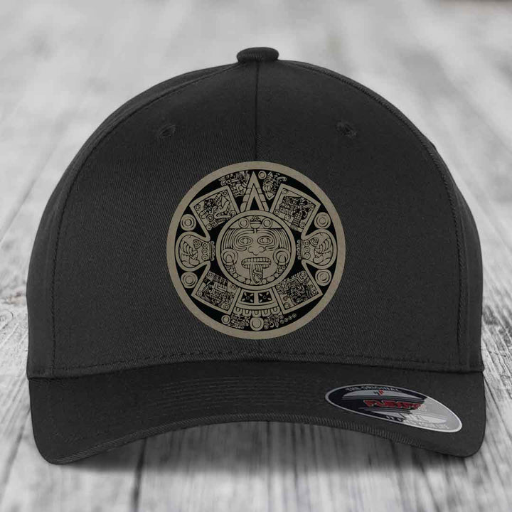 Black baseball cap with Aztec calendar patch on front, sitting on wooden surface.
