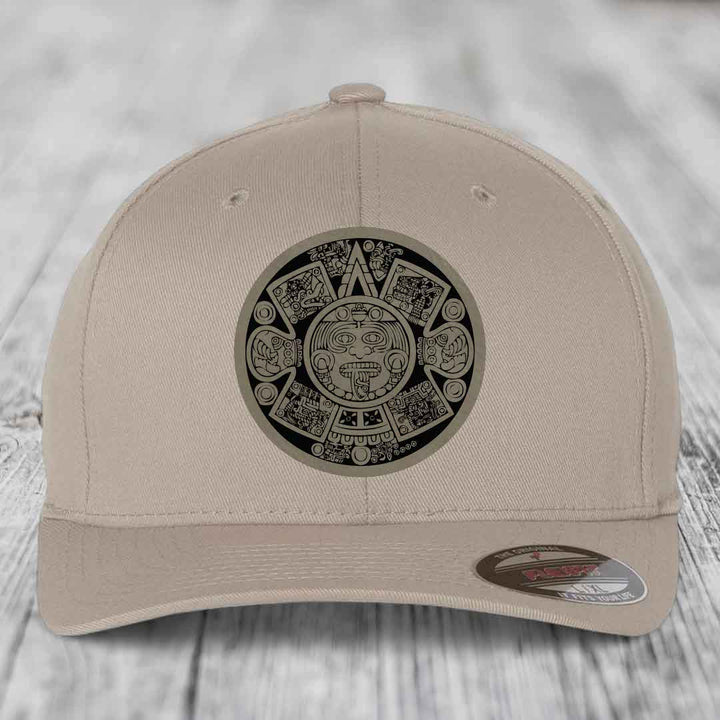 Beige baseball cap with Aztec calendar patch on front, displayed on wooden surface.
