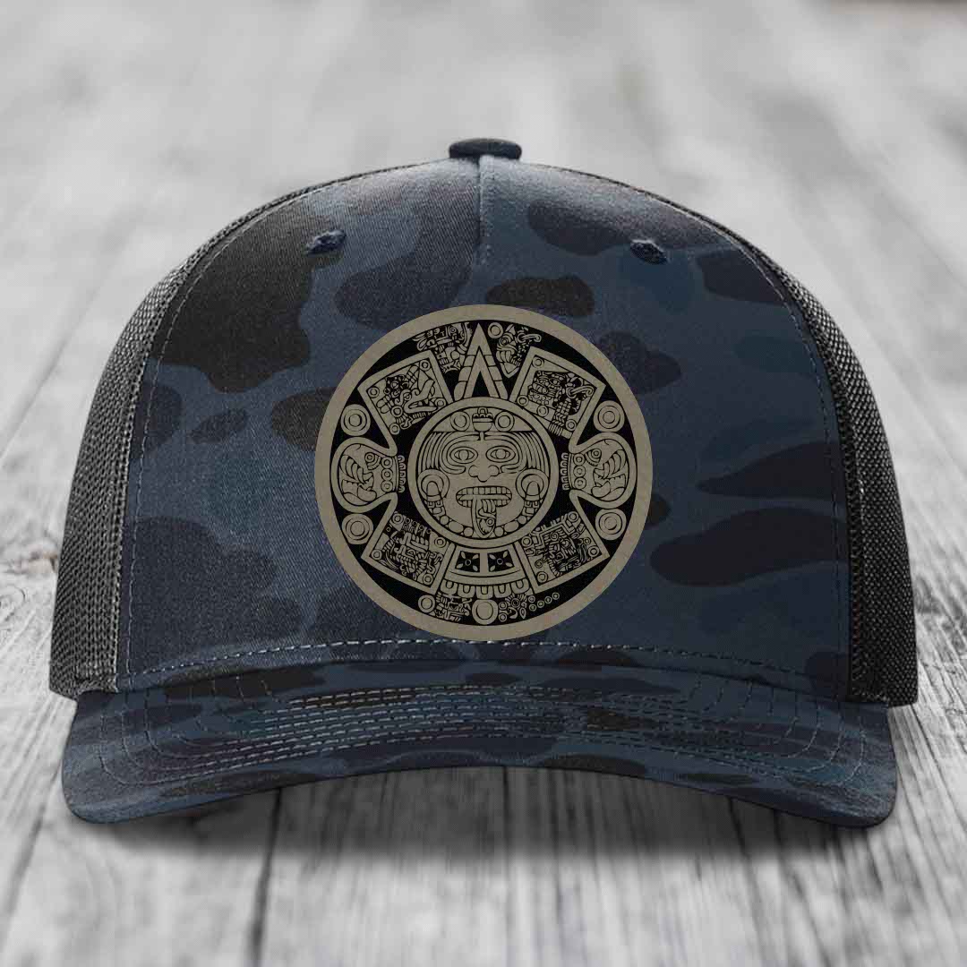 Camouflage trucker hat with Aztec calendar patch on front, placed on wooden surface.