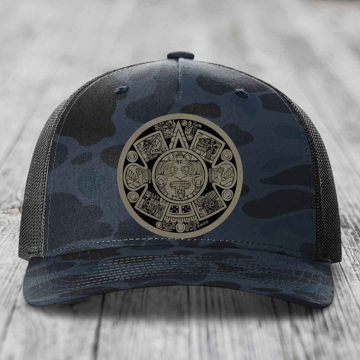 Camouflage trucker hat with Aztec calendar patch on front, placed on wooden surface.