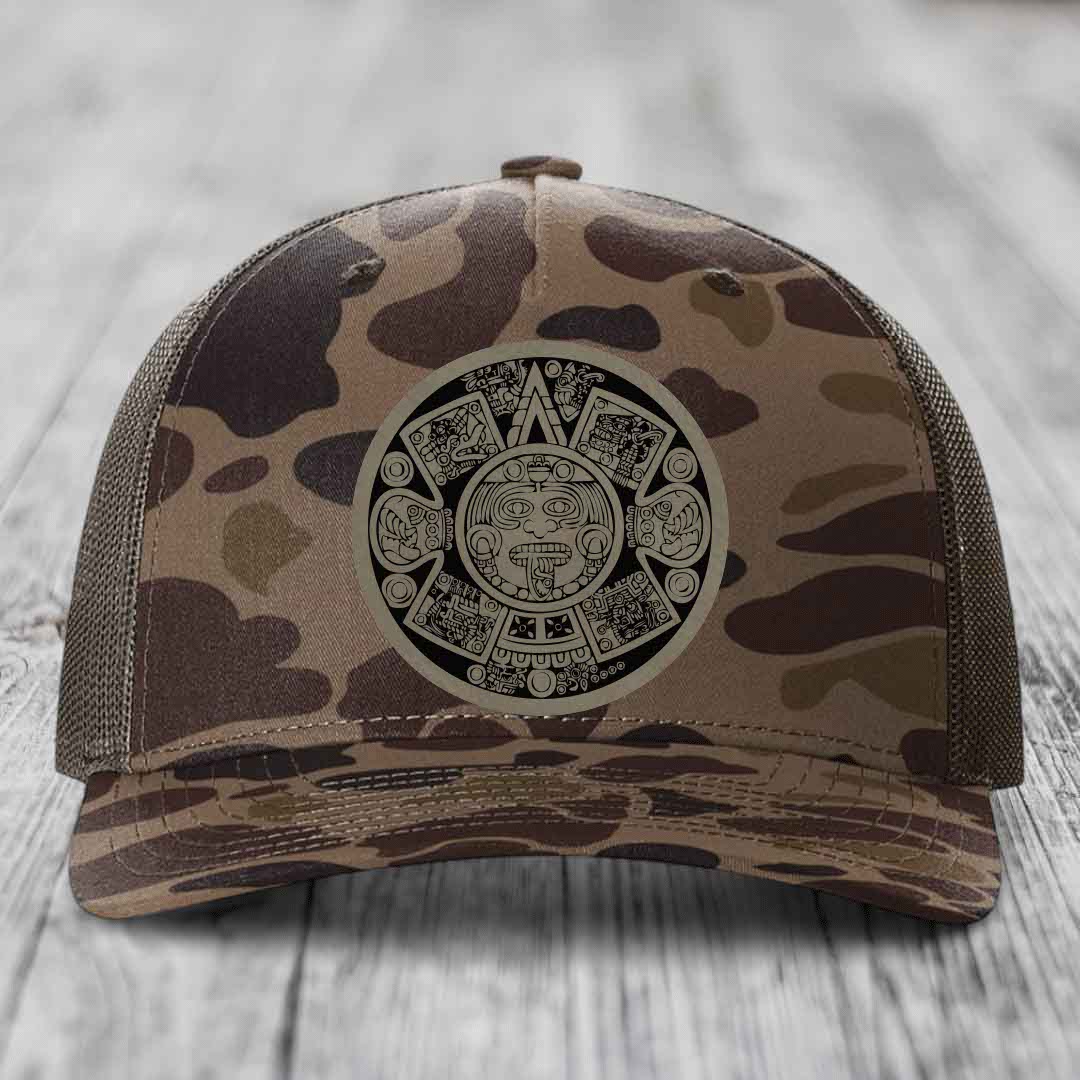 Camouflage trucker hat with Aztec calendar patch on front, placed on wooden surface.