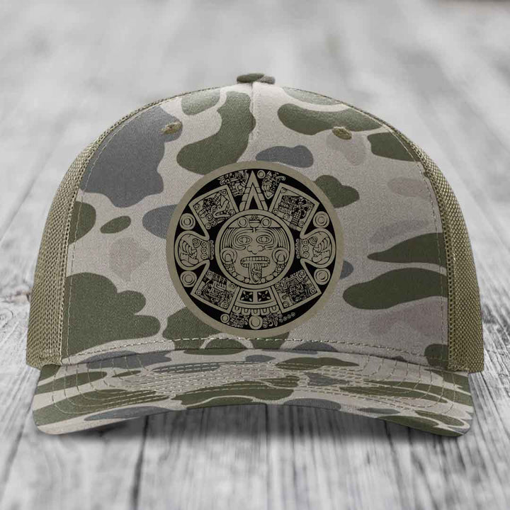 Camouflage trucker hat with Aztec calendar patch on front, placed on wooden surface.