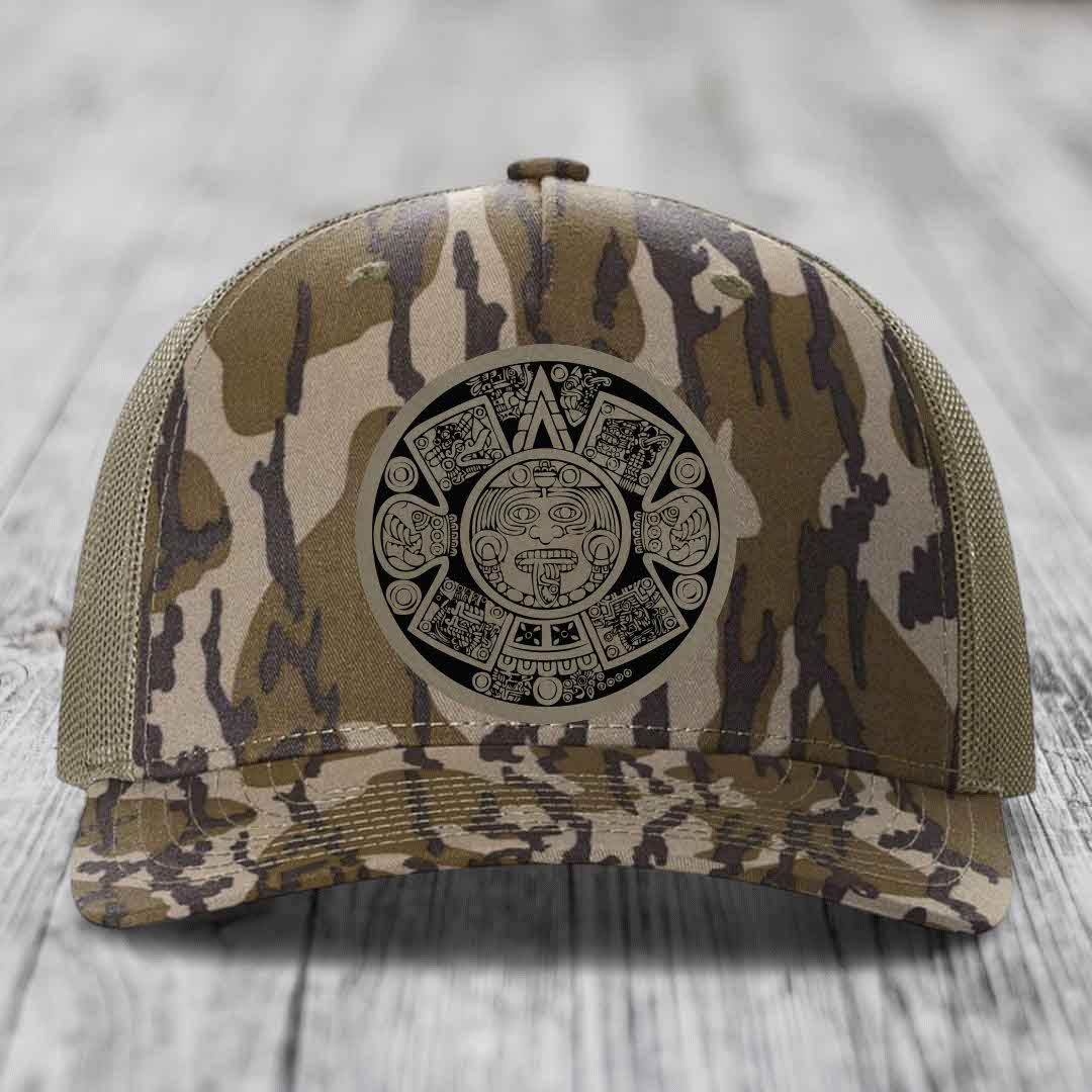 Camouflage trucker hat with Aztec calendar patch on front, placed on wooden surface.