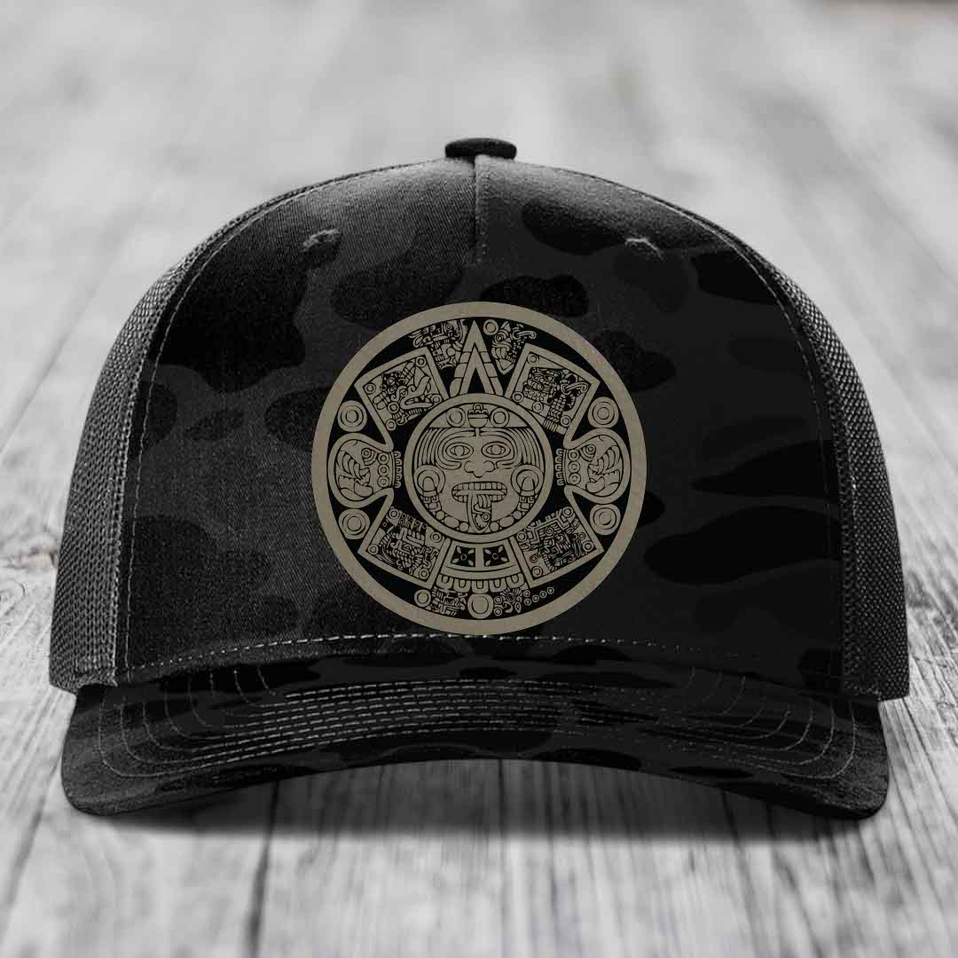 Black trucker hat with Aztec calendar design on front panel, placed on wooden surface.