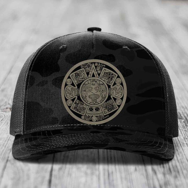 Black trucker hat with Aztec calendar design on front panel, placed on wooden surface.