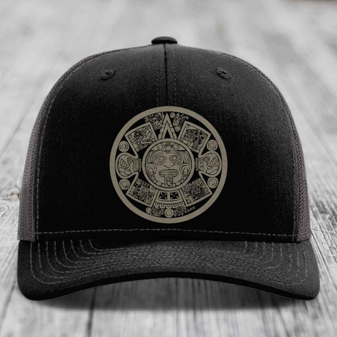 Black trucker hat with Aztec calendar design on front, displayed on wooden surface.